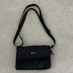 Svart axelväska i skinn med ringdetaljer  - Snygg svart axelväska från Zebra i skinn med justerbar rem och coola ringdetaljer på framsidan. Väskan har flera fack med dragkedjor. Pris kan diskuteras!