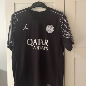 PSG svart matchtröja D. Doué #14 - Säljer en svart PSG fotbollströja med vita detaljer och tryck, Jordan-logga och Qatar Airways sponsor på bröstet. Nummer 14 och D. Doué på ryggen. Snygga grafiska linjer på ärmarna. Tillverkad i lätt och ventilerande material.