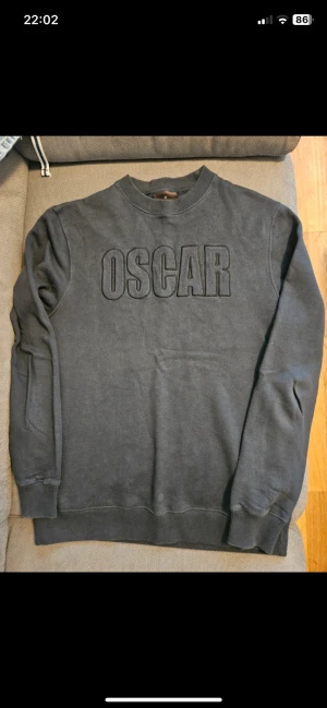 Mörkgrå sweatshirt Oscar Jacobson M - Snygg mörkgrå sweatshirt från Oscar Jacobson i storlek M. Tröjan har ett stort broderat OSCRAR-tryck på bröstet och klassisk rund hals. Mjukt material med ribbade muddar vid ärmslut och nederkant, perfekt för chill dagar. Sitter mer som S