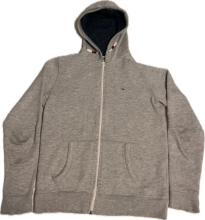 Grå hoodie från Hilfiger Denim - Snygg grå hoodie från Hilfiger Denim i storlek M, dock lite liten i storlek. Tröjan har hel dragkedja, huva med mörkblå insida och två praktiska fickor framtill. Liten broderad logga på bröstet och ribbade muddar vid ärmslut och nederkant. Perfekt för chill dagar.