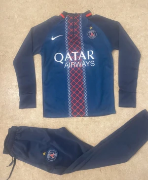 PSG träningsset Nike blå - Snyggt träningsset från Paris Saint-Germain med långärmad tröja och matchande byxor i mörkblått. Tröjan har PSG-logga, Nike-swoosh och Qatar Airways-tryck samt ett randigt mönster i rött och vitt framtill. Byxorna har dragsko och PSG-emblem. Perfekt för fotbollsträning.