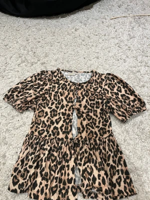 Leopardmönstrad kort klänning Gina Tricot - Kort klänning från Gina Tricot med leopardmönster i beige och svart. Modellen har puffiga korta ärmar, rund halsringning och en söt knytning framtill. Materialet är mjukt och lätt, perfekt för en trendig look.