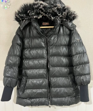 Svart pufferjacka med päls från Aysema - Svart pufferjacka från Aysema med fluffig huva och snyggt pälsdetalj i svart och vitt. Jackan har dragkedja framtill, ribbade muddar och två fickor med knapp. Perfekt för kalla dagar och har en cool, oversized vibe.