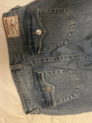 Bootcut jeans från True Religion - Snygga ljusblå jeans från True Religion med klassiska ficklock och silverfärgade knappar. Bootcut-modell med breda ben och coola kontrastsömmar. Jeansen har en faded effekt på framsidan och är tillverkade i jeansmaterial med bomullskänsla. Dock så har de hål mellan benen som alltid går att sy ihop 