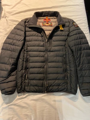 Svart pufferjacka från Parajumpers - Säljer en svart pufferjacka från Parajumpers med quiltad design och hög krage. Jackan har två sidofickor med dragkedja, emblem på ärmen och snygga detaljer i orange och gul vid halsen. Perfekt för kalla dagar och riktigt stilren.