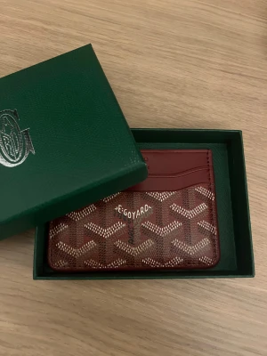 Röd Goyaard plånbok med mönster - Snygg och stilren plånbok från Goyaard i rött skinn med det klassiska Goyaard-mönstret i beige och vitt. Perfekt storlek för kort och cash, med flera fack och exklusiv vibe. Kommer i original grön ask med logga.