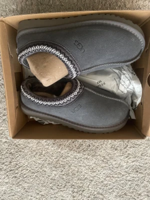 Grå UGG slip-on mockaskor - Säljer ett par grå slip-on skor från UGG med rund tå och platt sula. Skorna är tillverkade i mjuk mocka och har ett stickat, mönstrat band runt öppningen. Insidan är fodrad med päls för extra komfort och värme. Perfekta för kalla dagar och chill hemma.