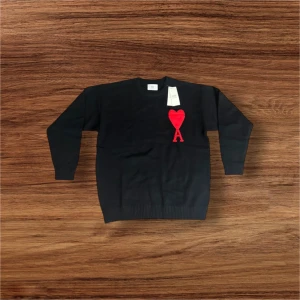 Svart stickad tröja från Ami - Svart stickad tröja från Ami Alexandre Mattiussi med ett stort rött hjärta och bokstaven A broderat på bröstet. Tröjan har rund hals, ribbade muddar och lång ärm. Perfekt för dig som gillar stilrena detaljer och vill sticka ut med en ikonisk design.