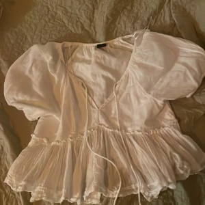 Vit blus med puffärm från Gina Tricot - Supersöt vit blus från Gina Tricot i storlek S. Blusen har puffiga korta ärmar, v-ringning med snörning och volangkant nedtill. Materialet är lätt och luftigt, perfekt för varma dagar. Snygg att styla med jeans eller kjol för en trendig look.