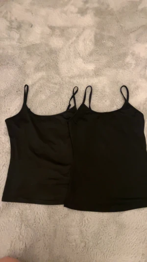 Svarta linnetoppar med smala axelband - Två svarta linnetoppar med smala axelband säljes. De har en enkel och stilren design med rundad halsringning och ärmlös passform. Perfekta att styla med jeans eller kjol för en clean look. Materialet känns mjukt och följsamt mot huden.