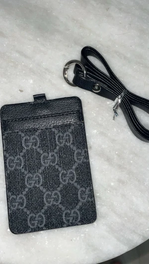 Svart plånbok med mönster - Snygg svart plånbok med klassiskt monogram mönster i grått. Kommer med en matchande svart rem och metallring. Perfekt för kort och småsaker, stilren och enkel att bära runt halsen eller i väskan.