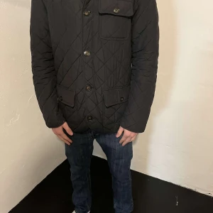 Svart quiltad jacka från Gant - Svart quiltad jacka från Gant med snygga knappar och fickor framtill. Jackan har förstärkta armbågslappar i mörkare tyg och ett klassiskt rutmönster över hela ytan. Perfekt för dig som gillar stilren och tidlös design.