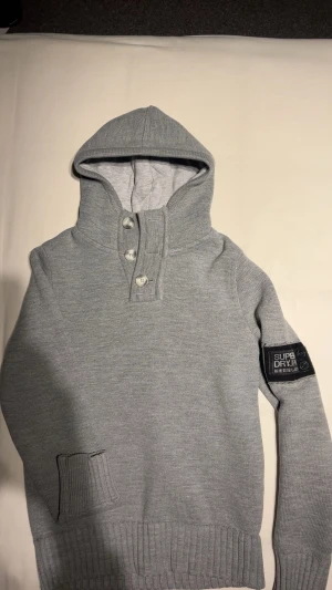 Grå hoodie från Superdry  - Snygg grå hoodie från Superdry med ribbade muddar och huva. Tröjan har tre knappar vid halsen och ett svart Superdry-märke på ärmen. Materialet är stickat och ger en skön känsla. Perfekt för dig som gillar stilren och enkel design. Storlek Medium.