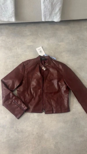 Mörkbrun skinnjacka med dragkedja - Snygg mörkbrun skinnjacka från C&A med raka linjer och cool ståkrage. Jackan har dragkedja framtill, två knappar vid halsen och ficka med dragkedja på bröstet. Perfekt för dig som gillar en stilren och edgy look.