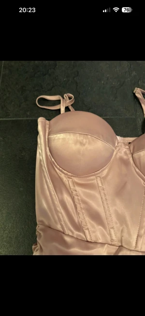 Rosafärgad satin klänning - Snygg kort klänning i rosafärgad satin med figurnära passform och justerbara axelband. Klänningen har markerade kupor och sömmar framtill som ger en bustier-look. Perfekt för dig som vill ha en elegant och trendig stil.