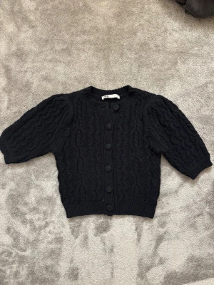 Marinblå croppad kofta från Zara - Marinblå stickad kofta från Zara i storlek S. Croppad modell med korta ärmar och snyggt kabelstickat mönster. Rund halsringning och knappar framtill. Perfekt att styla med jeans eller kjol för en trendig look. 