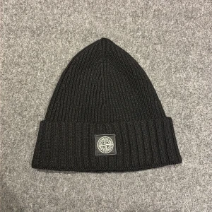 Svart ribbstickad mössa Stone Island - Svart ribbstickad mössa från Stone Island med klassisk logopatch framtill. Mössan är i mjuk ull och har en bred uppvikt kant för extra stilpoäng. Perfekt accessoar för att hålla värmen och lyfta din outfit under kyliga dagar.