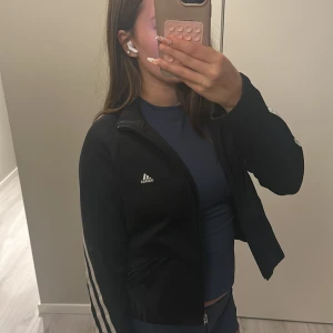 Svart Adidas gym kofta med vita ränder - Säljer en svart Adidas gym kofta med kla vita ränder längs ärmarna och logga på bröstet. 