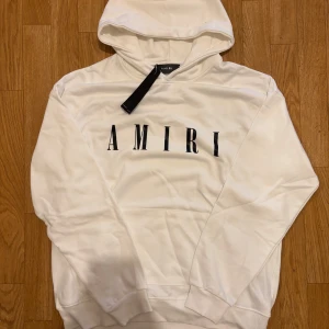 Vit Amiri hoodie - Säljer en stilren vit hoodie från Amiri med svart logotyptryck på bröstet. Tröjan har huva, långa ärmar och ribbade muddar. Kvaliten är på topp och den är helt oanvänd. Skriv om du har några frågor:)