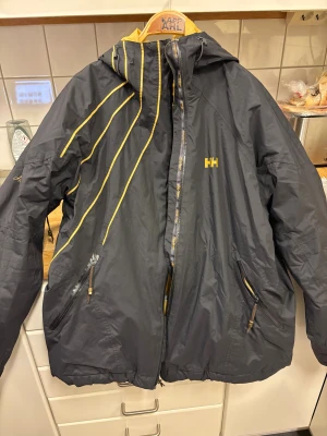 Svart och gul vindjacka Helly Hansen XL - Svart vindjacka från Helly Hansen i storlek XL med gul insida och detaljer. Jackan har huva, dragkedja framtill och två fickor med dragkedja. Snygga gula linjer på framsidan och HH-logga på bröstet och insidan. Perfekt för dig som gillar sportig stil.