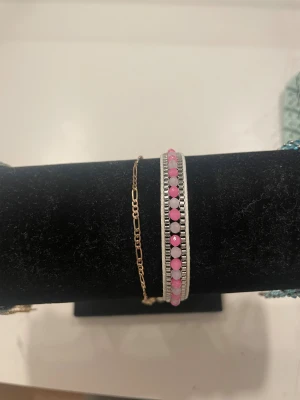 Två armband i guld och silver med rosa pärlor - Säljer två snygga armband, ett tunt guldarmband med kedjedetaljer och ett bredare silverfärgat armband med rad av rosa och ljuslila pärlor. Perfekt för dig som gillar att mixa olika stilar och färger.