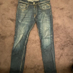 Replay Anbass jeans blå slim fit - Snygga Replay Anbass jeans i blå tvätt med slitningar och kontrastsömmar. Slim fit-modell med klassiska fem fickor, dragkedja och läderpatch bak. Ikonisk Replay-logga på bakfickan och detaljerad sömnad framtill. Perfekta för dig som gillar en modern och stilren look. Storlek är 28/30! 