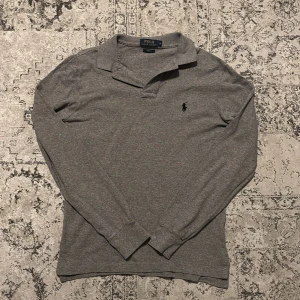 Grå långärmad pikétröja Polo Ralph Lauren - Snygg grå långärmad pikétröja från Polo Ralph Lauren i slim fit. Klassisk krage och svart broderad logga på bröstet. Mjuk bomullskvalitet och ribbade muddar vid ärmslut. Perfekt för dig som gillar stilrena och tidlösa plagg.