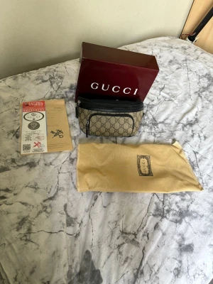 GG väska - Säljer en beige axelväska med klassiskt GG-mönster i mörkbrunt och svarta detaljer. Väskan har justerbart svart band, dragkedja och en extra ficka framtill. Insidan är ljusbeige och mjuk. Kommer med originalförpackning och dustbag.