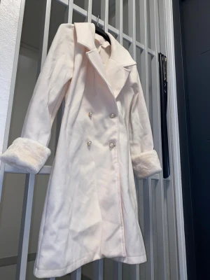 Vit kappa med fuskpäls och pärlknappar - Snygg vit kappa med bred krage och fluffiga fuskpälsdetaljer på ärmsluten. Dubbelknäppt framtill med pärlknappar som ger en lyxig vibe. Perfekt för dig som vill ha en elegant och mysig look under kyliga dagar.