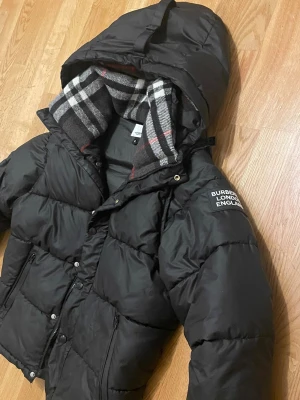 Svart dunväst från Burberry - Svart dunväst från Burberry med huva och klassiskt rutigt foder i grått, vitt och rött. Västen har dragkedja och tryckknappar framtill samt två fickor med dragkedja. Märkeslogga på vänster axel. Perfekt för kyliga dagar.