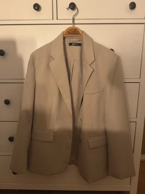 Beige kavaj från Gina Tricot - Stilren beige kavaj från Gina Tricot med klassisk krage och två fickor med lock framtill. Kavajen har en rak passform och stängs med knappar. Perfekt för en clean och modern look.