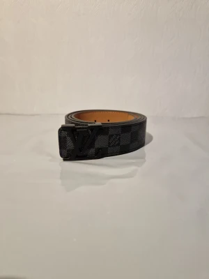 Louis Vuitton rutigt svart bälte - Snyggt svart bälte från Louis Vuitton med klassiskt rutigt mönster och stor LV-logga på spännet. Bältet är gjort i skinn och har en stilren design som sticker ut. Perfekt accessoar för att lyfta din outfit.