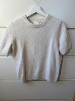 Ljusbeige stickad tröja från H&M - Mysig ljusbeige stickad tröja från H&M i storlek S. Tröjan har korta ärmar, rund hals och ribbade detaljer vid hals, ärmslut och nederkant. 