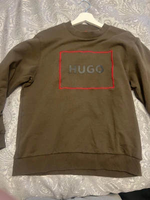 Brun sweatshirt från Hugo - Snygg brun sweatshirt från Hugo med grått tryck på bröstet och en röd markerad ruta runt loggan. Tröjan har rund hals, ribbade muddar och är långärmad. Perfekt för dig som gillar stilrena och moderna plagg med tydlig branding.