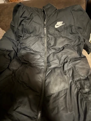 Svart pufferjacka från Nike  - Svart pufferjacka från Nike i storlek XL, med klassisk Nike-logga på bröstet och hel dragkedja framtill. Jackan har en enkel, sportig design och är tillverkad i syntetmaterial som håller värmen. Perfekt för kalla dagar och streetwear-stil.
