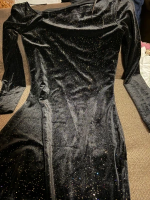 Svart glittrig sammetsklänning - Supersnygg svart sammetsklänning med glittriga detaljer över hela tyget. Klänningen har lång ärm och en figurnära passform som ger en elegant siluett. Perfekt för dig som vill sticka ut med lite extra glitter.