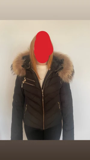 Svart pufferjacka Hollies med päls - Snygg svart pufferjacka från Hollies med fluffig beige päls på huvan. Jackan har guldfärgade dragkedjor, flera fickor och ett rundat Hollies-märke på ärmen. Perfekt för kalla dagar och har en riktigt schysst siluett med quiltade detaljer. Riktig päls. Den är inte sönder någonstans!!