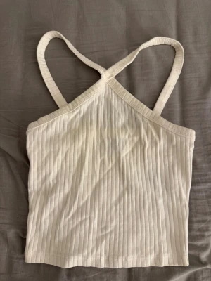 Ribbad vit linnetopp med korsade band - Säljer en fräsch ribbad linnetopp i vit färg med korsade axelband. Toppen är ärmlös och har en snygg croppad passform som passar perfekt till jeans eller kjol. Materialet är mjukt och stretchigt, troligen bomull. Enkel och stilren design för en clean look.