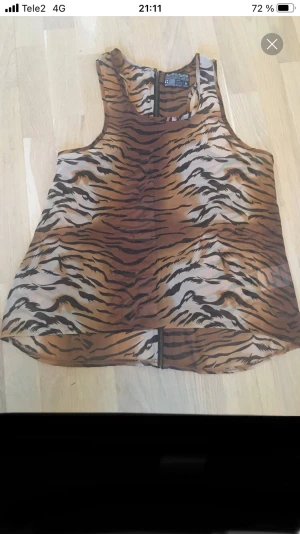 Tigerprintad ärmlös linnetopp Saints & Mortals - Säljer en cool ärmlös linnetopp från Saints & Mortals i transparent syntetmaterial med tigerprint i brunt, beige och svart. Toppen har rund hals och dragkedja längs hela ryggen för extra edge. Perfekt för dig som gillar djurmönster och vill sticka ut.