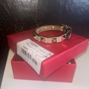 Valentino armband med nitar - Coolt armband från Valentino i beige skinn med ikoniska pyramidformade nitar i guld och silver. Justerbart spänne och snygg kontrastkant. Perfekt accessoar för att lyfta din outfit.