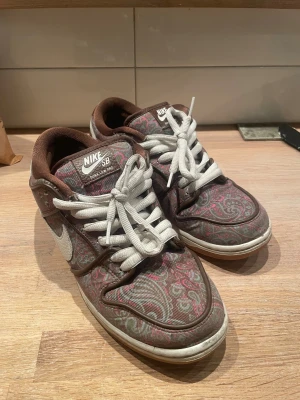 Nike SB Dunk Low Pro Paisley 41 - Nike SB Dunk Low Pro med paisleymönster i brunt, rosa och grått. Skorna har vit swoosh, vita snören och gummisula med rosa detalj undertill. Ovandelen är i textil med tydligt paisleyprint och bruna detaljer runt häl och plös. Perfekt för dig som gillar unika sneakers.