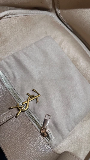 Beige axelväska från Saint Laurent - Snygg beige axelväska från Saint Laurent med klassisk YSL-logga i guld på framsidan. Väskan är tillverkad i strukturerat skinn och har en bred axelrem. Insidan har en praktisk ficka med dragkedja och mjukt foder. Perfekt för dig som gillar stilrena detaljer.