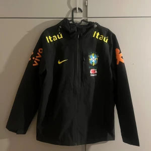 Svart Brasil Nike vindjacka L - Svart vindjacka från Nike med Brasil CBF-logga och sponsormärken i gult och orange. Jackan har huva, hel dragkedja och tryck på både bröst och ärmar. Perfekt för dig som gillar fotboll och vill ha en sportig look.