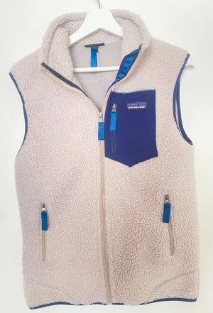 Beige fleeceväst från Patagonia - Mysig beige fleeceväst från Patagonia med blå detaljer och dragkedjor. Västen har hög krage, tre fickor med dragkedja och en blå bröstficka med logga. Perfekt för lager-på-lager och chill dagar.