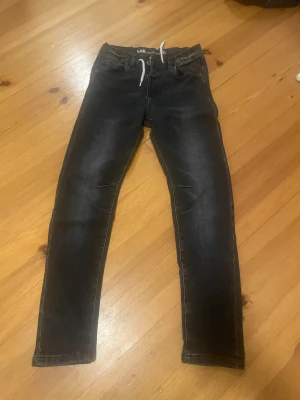 Svarta jeansbyxor från LAB Industries - Snygga svarta jeansbyxor från LAB Industries med vit snörning i midjan. Jeansen har en smal passform och är tillverkade i ett hållbart denim-material. Perfekta för dig som gillar en modern och stilren look.