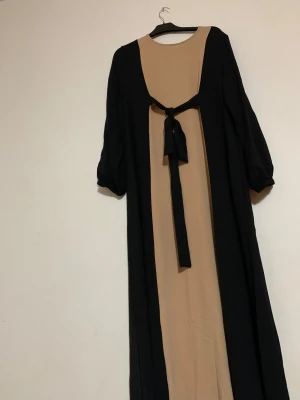 Beige och svart långklänning med knyt - Snygg långklänning i beige och svart med rund hals och långa ärmar. Klänningen har en cool kontrast mellan färgerna och ett knytband i ryggen som ger extra stil. Materialet känns mjukt och har ett fint fall. Perfekt för dig som gillar en unik look.