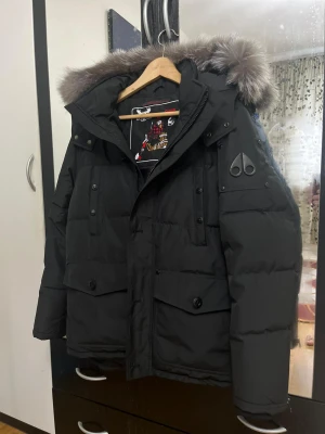 Svart dunjacka från Moose Knuckles onyx - Säljer en svart dunjacka från Moose Knuckles. Sällsynt model som är slutsåld överallt. Kommer med jätte fin silver pälskant på huvan och snygga logodetaljer på ärmen och framtill. Jackan har flera fickor med knappar och dragkedja, samt en schysst quiltad design som håller dig varm hela vintern.