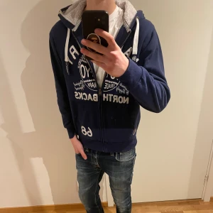 Mörkblå hoodie Tokyo Laundry - Mörkblå hoodie från Tokyo Laundry med vit textprint och dragkedja framtill. Utrustad med huva och vita snören, samt mjukt grått fleecefoder i luvan. Snygg streetstil med stora tryck och praktiska fickor.skriv vid funderingar🤝
