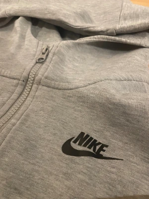 Grå Nike tech zip hoodie - Snygg grå zip hoodie från Nike i storlek 158-170cm. Tröjan har hel dragkedja, huva och svarta detaljer vid muddar och bröstet. Materialet är mjuk bomull och polyester, perfekt för chill eller träning. Nike-logga tryckt på bröstet och praktiska fickor framtill.