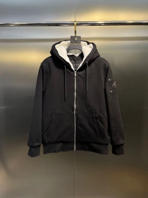 Svart teddyjacka från Moose Knuckles - Svart teddyjacka med vit fluffig huva från Moose Knuckles. Jackan har dragkedja framtill, snörning vid huvan, två stora fickor och en patch med logga på ärmen. Insidan är quiltad och fodrad för extra värme. Perfekt för dig som gillar streetwear och vill ha en snygg look.